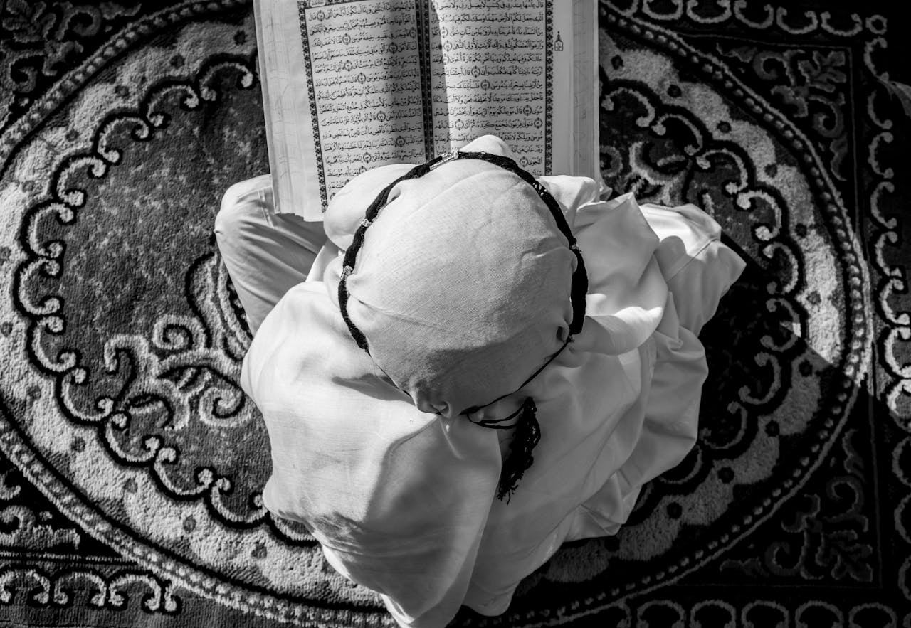 A child wearing traditional attire reads an open Quran on a patterned rug from above.